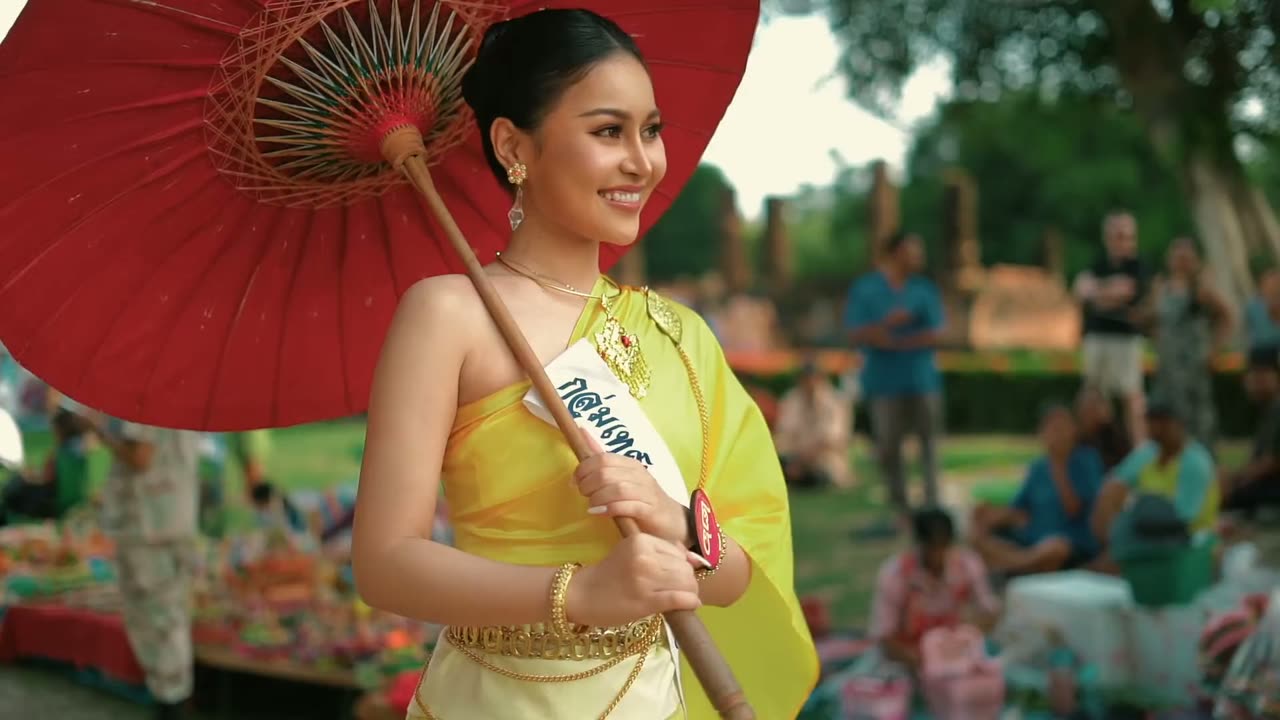 ผู้หญิงในชุดไทยสีเหลืองถือร่มแดง กลางลานจัดงานตอนกลางวัน